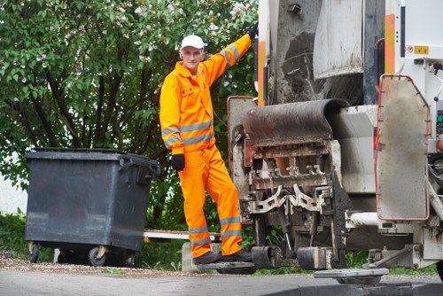 A waste collection worker wearing high-visibility orange safety clothing, including a jacket with reflective stripes and a white hard hat, stands on a paved surface next to a large commercial waste disposal truck. The worker is smiling and pointing towards the black wheeled waste bin positioned on the ground nearby, which is partially open with no contents visible. The waste truck is grey with an open rear compactor section, situated on the right side of the image, showing mechanical components and a partially visible conveyor belt used for crushing and lifting rubbish. Behind the scene, there are green leafy trees providing a natural backdrop, indicating an outdoor setting likely along a residential or commercial street. The scene captures the typical environment of rubbish collection in Thames Ditton, with clean, well-maintained equipment and a focus on efficient waste removal service by Commercial Waste Thames Ditton.