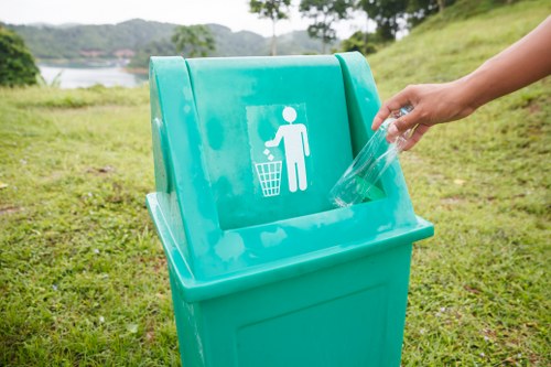 A person is seen discarding a clear plastic bottle into a bright turquoise outdoor rubbish bin with a white graphic of a person and a trash can symbol on the front, situated on a grassy area. The background features a scenic landscape with green trees, a body of water, and distant hills under a cloudy sky. The bin is positioned on a natural grass surface, and the person's hand is visible as they place the item into the bin, reflecting an act of waste disposal. This scene demonstrates an outdoor capacity for rubbish collection, consistent with waste management services provided by companies such as Commercial Waste Thames Ditton, serving local areas in Thames Ditton and surrounding regions.