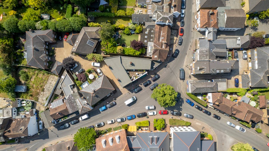 An aerial view of a residential street with numerous parked cars lining both sides of the narrow, curved road, surrounded by closely packed houses with varied roofing materials such as tiles and slate