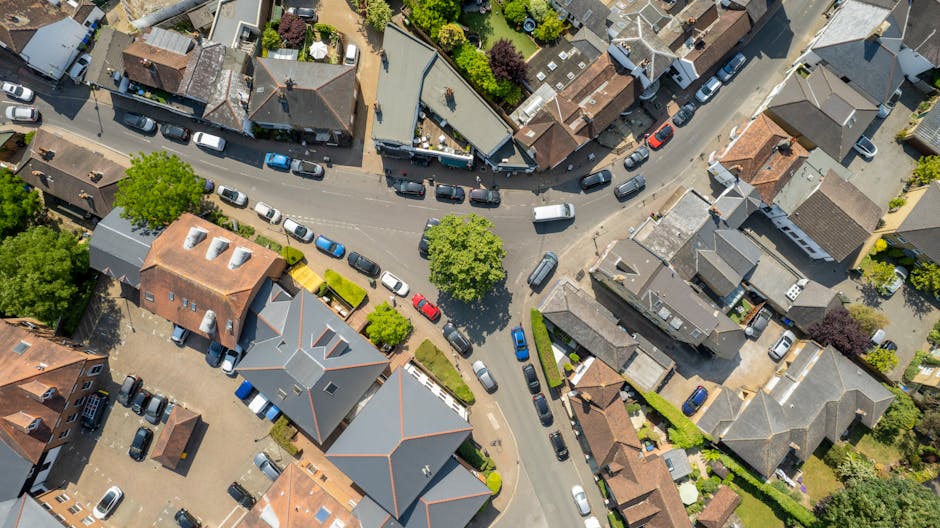 An aerial view of a busy street scene featuring a white, two-story commercial building with a flat roof and traditional sash windows, occupied by a small shop or café with an awning. The pavement in 