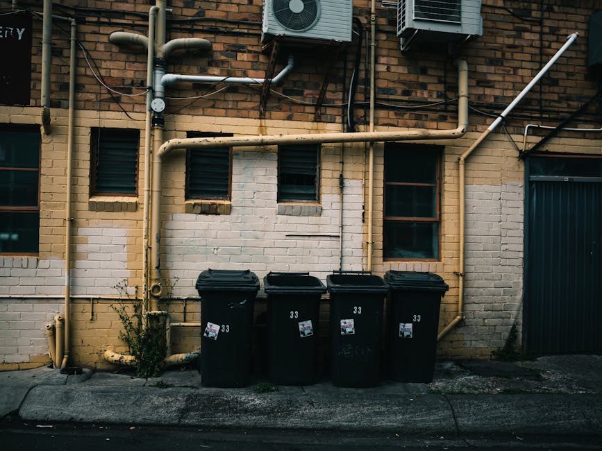 Four black wheeled waste bins with closed lids are positioned in a row against a façade of a brick building, situated directly on a concrete pavement curb. Each bin has a white label and a small stic