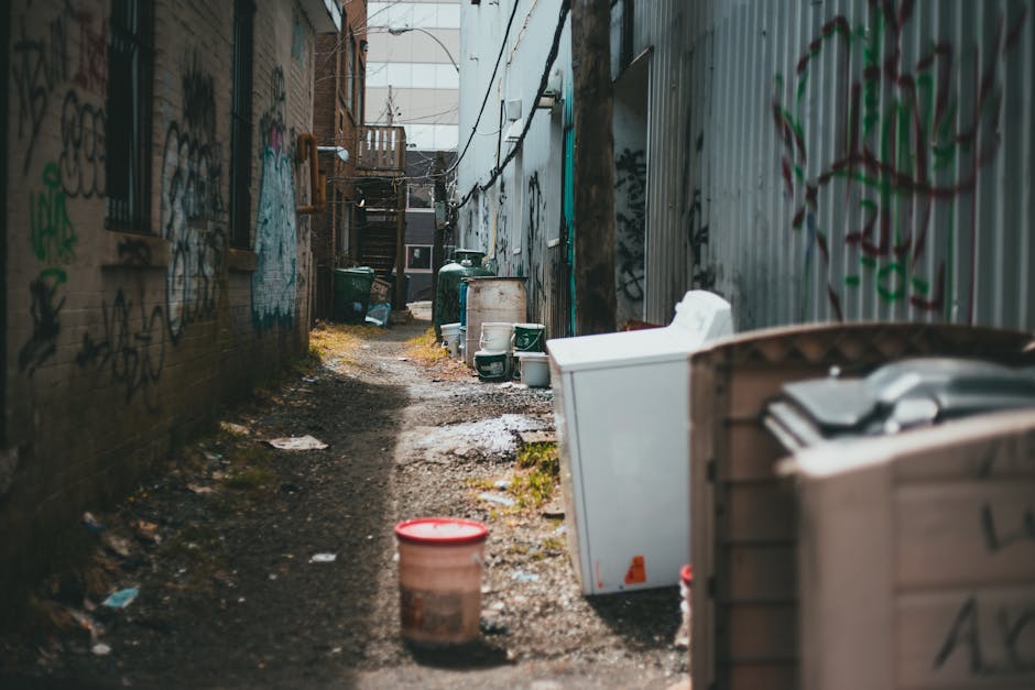 Four black wheeled waste bins with closed lids are positioned in a row against a façade of a brick building, situated directly on a concrete pavement curb. Each bin has a white label and a small stic